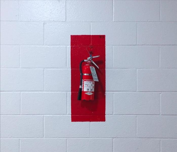Small fire extinguisher hanging on red rectangle on a white block wall.