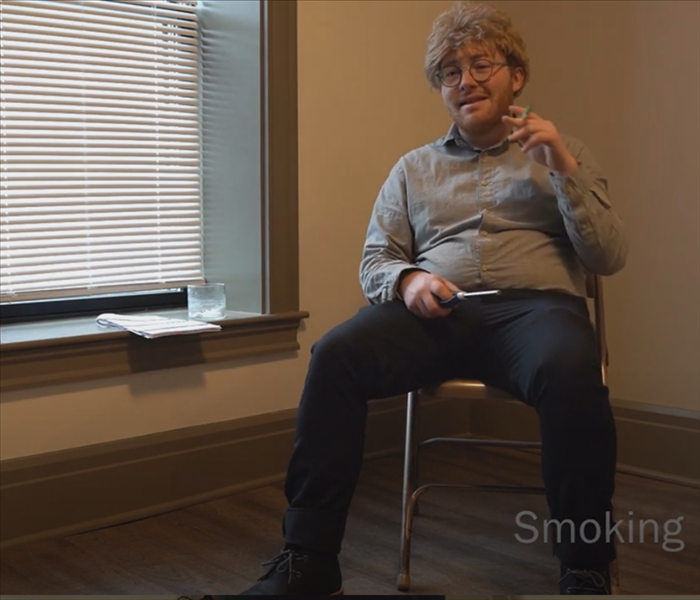 A man in a gray shirt and blonde wig pretending to smoke a pen in an empty apartment.